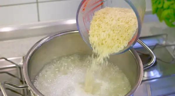 Reis im Topf kochen: Eine Hand schüttet Reis aus einem Messbecher in kochendes Wasser.