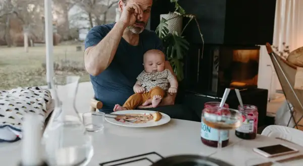 Trött pappa försöker äta frukost medan en bebis vaknar tidigt. Barnet sitter i pappans knä, en tallrik med mat står på bordet.