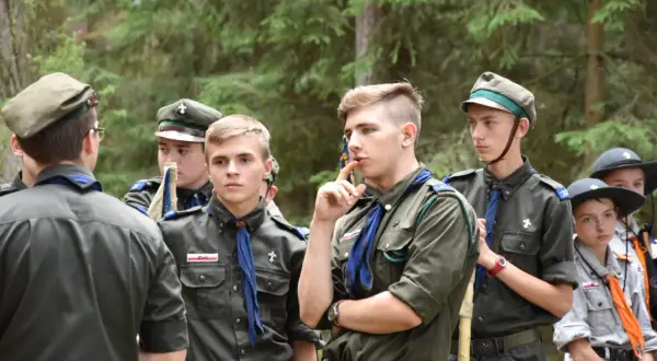 Harcerze w mundurach czekają na rozpoczęcie modlitwy obozowej. W lesie panuje skupienie.