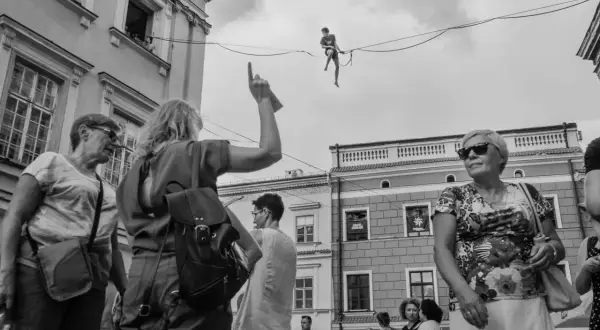 Street photography: widzowie patrzą na akrobatę na linie. Kobieta wskazuje palcem w górę.