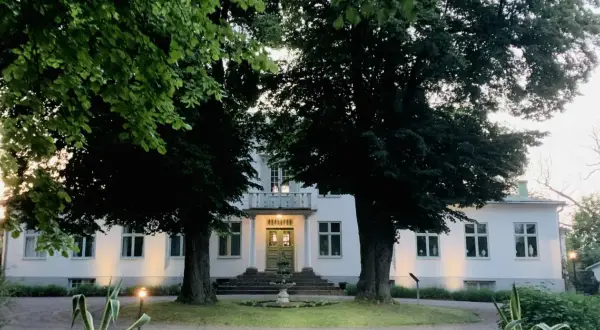 Vitt hus med gröna dörrar och fönster, omgivet av träd. En fontän framför entrén. Drottning Victorias vilohem.