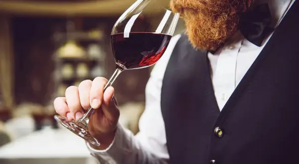 Uomo con barba rossa assapora un bicchiere di vino rosso durante una degustazione.