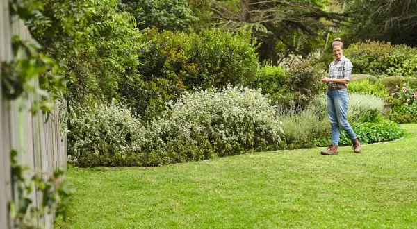 Mujer sonríe en un jardín exuberante, disfrutando del cuidado del césped en primavera. El césped verde y los arbustos floridos crean un paisaje idílico.