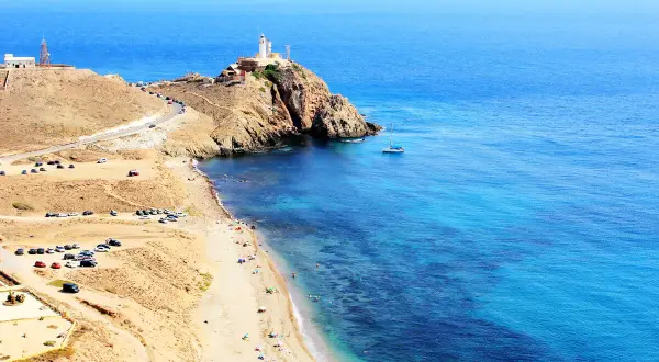 Costa de **Cabo de Gata** con faro en la cima de un acantilado, playa con gente y aguas cristalinas.
