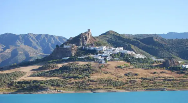 Pueblo blanco en la cima de una colina rocosa, con un castillo en ruinas. Un lugar ideal que ver cerca de Ronda, con un embalse azul en primer plano.