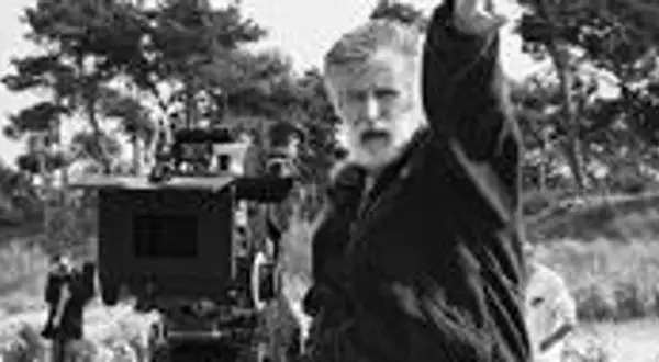 Edward Kłosiński, z siwą brodą, gestykuluje na planie filmowym. W tle widać kamerę i drzewa.