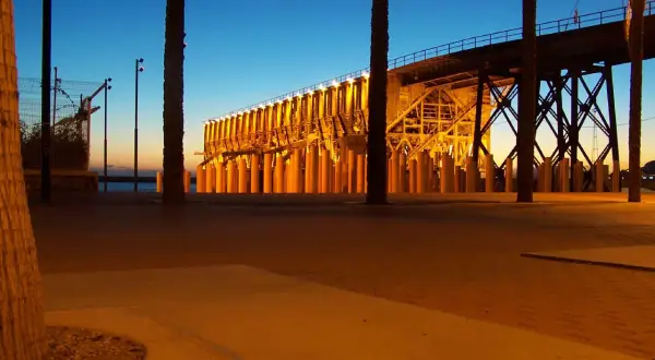Atardecer en el parque de las almadrabillas, con palmeras y un puente iluminado sobre el mar.