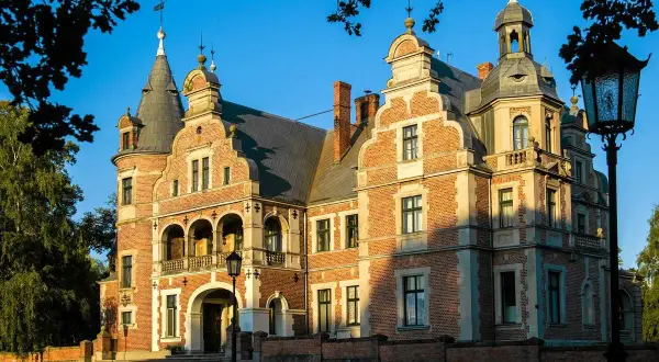 Pałac Kobylniki, okazała budowla z cegły z wieżyczkami i arkadowym gankiem, lśni w słońcu.