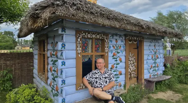 Mężczyzna siedzi na ławce przed malowaną chatką w stylu wsi Zalipie. Chata ozdobiona jest kwiatowymi wzorami.