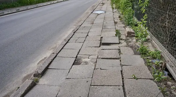 Zniszczony chodnik obok drogi. Płyty chodnikowe są popękane i nierówne, tworząc niebezpieczną ścieżkę dla pieszych.