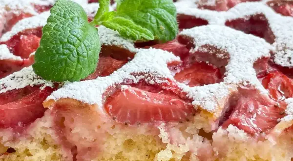 Puszyste ciasto jogurtowe z kawałkami truskawek, posypane cukrem pudrem i ozdobione listkiem mięty.