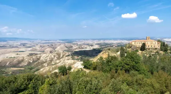 Panoramica delle **balze di Volterra**, con un casale in pietra in primo piano e dolci colline ondulate all'orizzonte sotto un cielo azzurro.