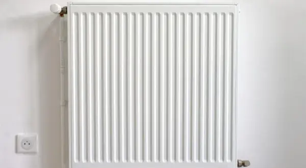 Biały kaloryfer żeberkowy na białej ścianie, z widocznym termostatem i gniazdkiem elektrycznym.