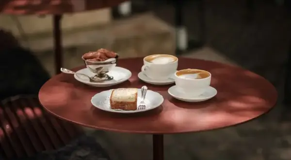 Två kaffekoppar med latte macchiato och en latte står på ett rött bord. Bredvid finns en bit kaka och en skål med chokladmousse.