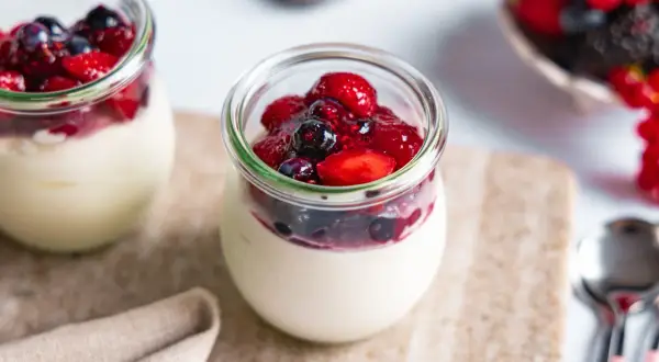 Zwei Gläser mit cremigem Dessert, gekrönt von Beeren und Fruchtsauce. Ein köstliches joghurtbombe rezept für den Sommer.