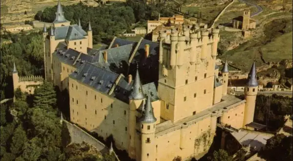 Vista aérea del Alcázar de Segovia, con sus torres y tejados, rodeado de vegetación. La sala de los reyes se intuye en su imponente estructura.