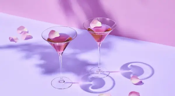 Dwa eleganckie drinki z martini rosato, ozdobione płatkami róż, unoszą się w delikatnym fioletowym świetle.
