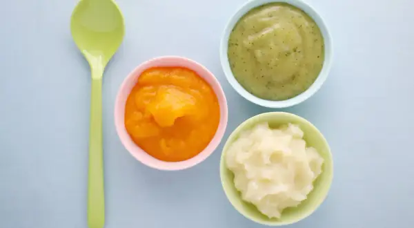 Drei Schälchen mit Brei (orange, grün, weiß) und ein Löffel. Ideal für die Beikosteinführung: ab wann Obstbrei Baby?