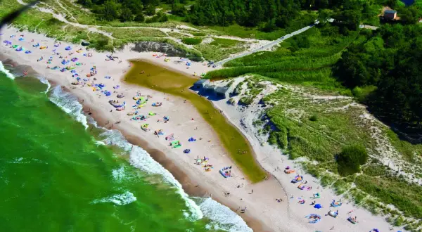 Plaża w Bobolinie tętni życiem. Ludzie odpoczywają na piasku, kąpią się w morzu i spacerują. W oddali widać las i malowniczą wydmę.