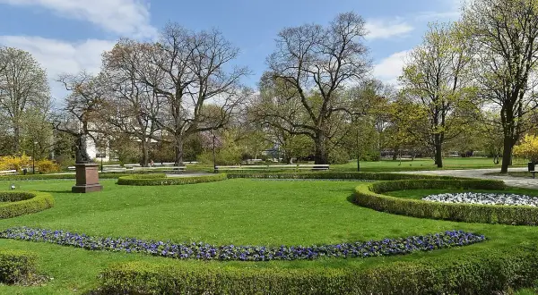 Piękny park miejski z rzeźbą, kwitnącymi rabatami i zielonymi trawnikami.
