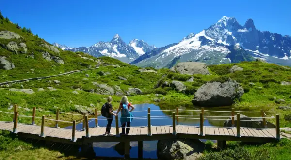 Zwei Wanderinnen auf einer Holzbrücke mit Blick auf den schneebedeckten Mont Blanc und eine grüne Berglandschaft.