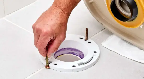 A hand tightens a bolt on a toilet flange installed on a tiled floor, preparing for a new toilet.