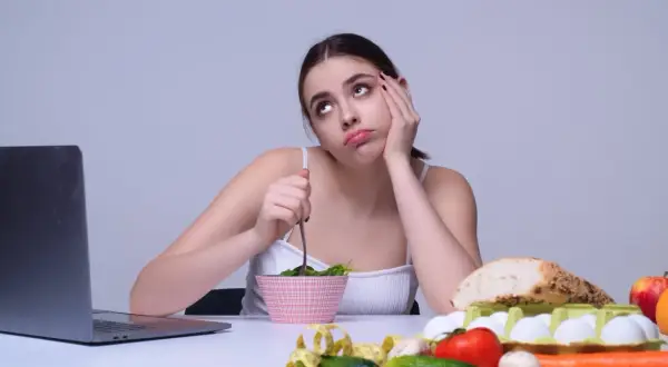 Une jeune femme s'ennuie en mangeant une salade, entourée de nourriture saine. Elle semble regretter de manger trop de fibres.