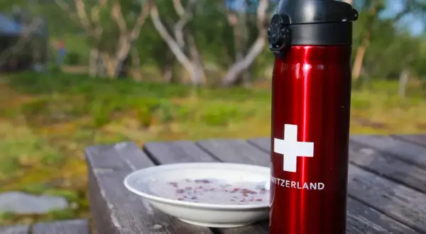 Röd termos med schweizisk flagga och en skål gröt på ett träbord. Perfekt för en frukost vandring i naturen.