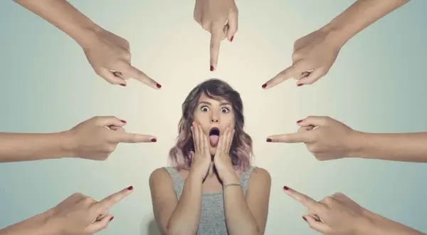Una ragazza con espressione spaventata, circondata da dita puntate. La sua bocca è spalancata, le mani sulle guance, simbolo della paura del giudizio altrui.