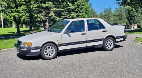 Vit Saab 9000 CD parkerad på en asfalterad väg med gröna träd och en blå himmel i bakgrunden.
