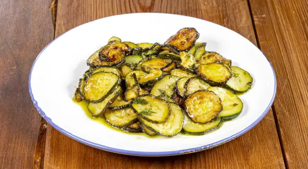 Piatto di zucchine alla poverella, fette sottili e dorate condite con olio e erbe aromatiche, su un piatto bianco con bordo blu.
