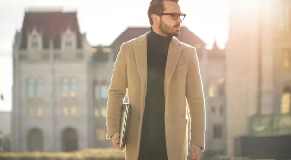 Mann im eleganten Mantel und Rollkragenpullover, mit Laptop und Koffer, verkörpert den "old money style" vor historischer Kulisse.