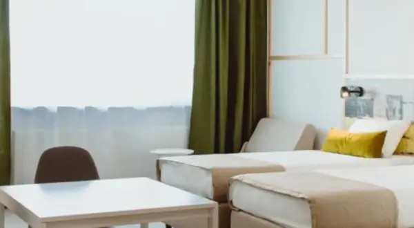 Pokój hotelowy z dwoma łóżkami, stołem i krzesłami, zielonymi zasłonami.