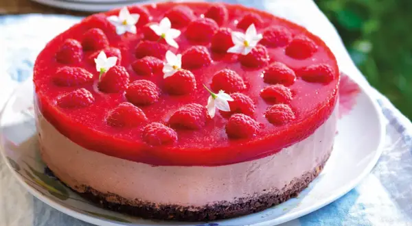 Cheesecake ricetta semplice con lamponi freschi e gelatina rossa, decorata con piccoli fiori bianchi.