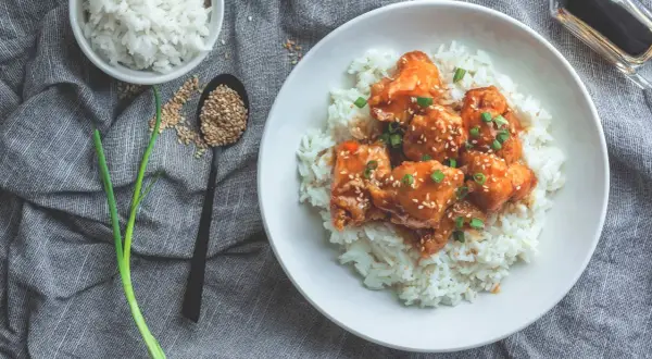 Ein Teller mit Reis und Hähnchen in süß-saurer Sauce, bestreut mit Sesam und Frühlingszwiebeln. Dieses Gericht hat Reis, Proteine und ist ein Genuss.