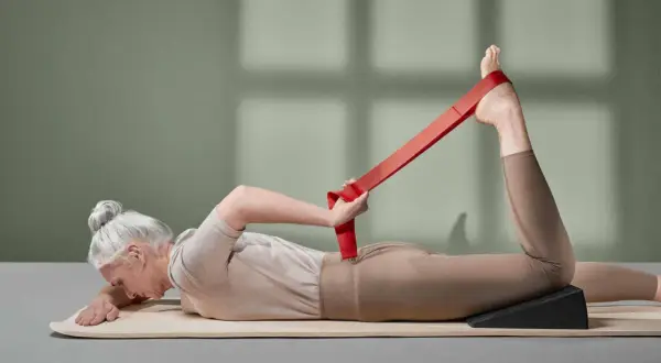 Ältere Frau mit grauem Dutt macht eine Dehnübung mit einem roten Band für das Bein.