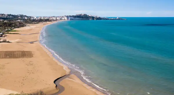 La spiaggia della Scialara si estende con sabbia dorata e mare azzurro, con il borgo bianco arroccato sulla scogliera in lontananza.