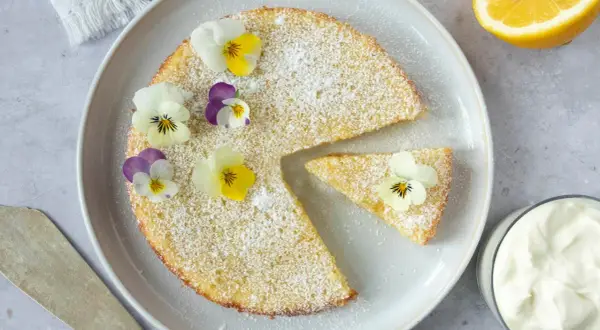 En saftig citron kladdkaka pudrad med florsocker och dekorerad med violer. En bit är uppskuren och en skål med grädde syns bredvid.