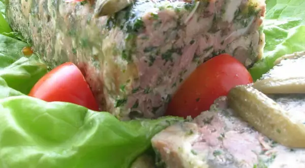 Tranche de pâté de tête maison, garnie de persil, servie sur des feuilles de laitue avec des tomates et des cornichons. Un délice pour une recette de pâté de tête.
