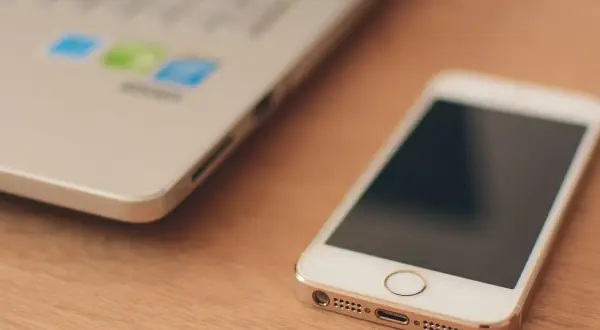 Biały smartfon leży obok laptopa. W tle widać logo z kolorowymi ikonami, sugerującymi uruchomione aplikacje w tle.