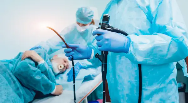 Lekarz w niebieskich rękawiczkach trzyma endoskop, przygotowując się do gastroskopii. Pacjent leży w tle.