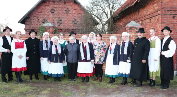 Grupa osób w tradycyjnych strojach wielkopolskich pozuje przed ceglanym budynkiem.
