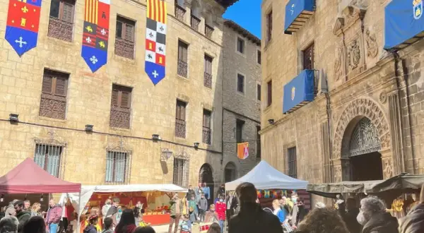 Gente disfruta del mercado medieval de las tres culturas, con puestos y banderas históricas adornando edificios antiguos.