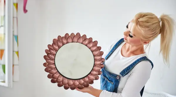 Okrągłe lustro w ramie z metalowych łyżeczek, jak zrobić je samemu. Kobieta w ogrodniczkach prezentuje efekt DIY.