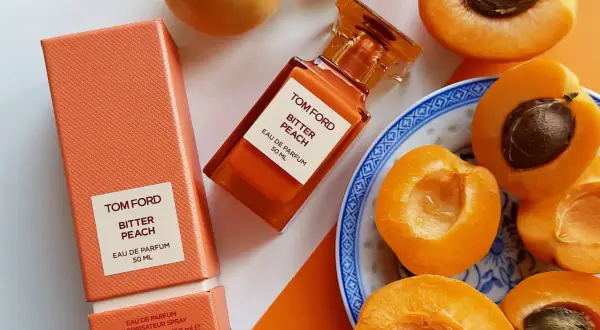 Pomarańczowe opakowanie i buteleczka perfum Tom Ford Bitter Peach otoczone soczystymi brzoskwiniami.
