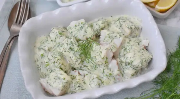 Pyszna ryba w sosie koperkowym, podana w białej misie, z dodatkiem cytryny i świeżego koperku.