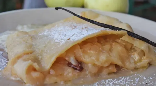 Ein Stück bayerischer Apfelstrudel, bestäubt mit Puderzucker und garniert mit einer Vanilleschote, daneben zwei Äpfel.