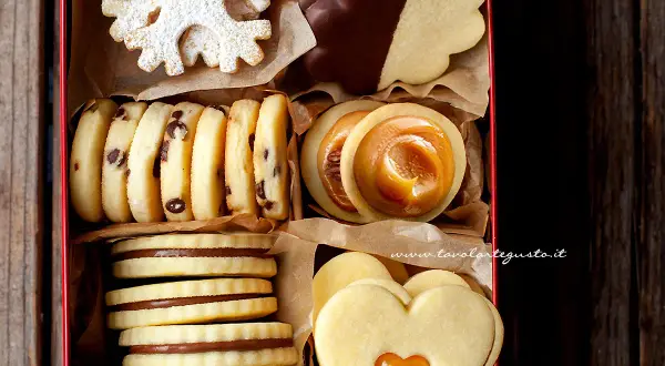 Scatola piena di biscotti al burro: fiocchi di neve, con gocce di cioccolato, ripieni di caramello, a forma di cuore e ricoperti di cioccolato.