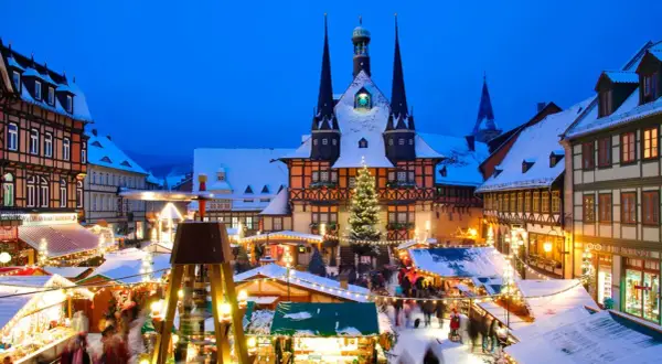 Ein verschneiter Weihnachtsmarkt in Deutschland mit beleuchteten Buden, einem Weihnachtsbaum und einem historischen Rathaus im Hintergrund.
