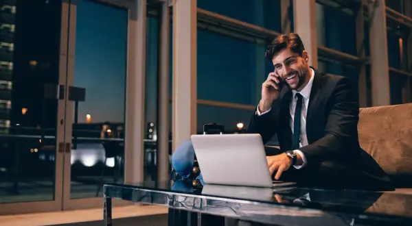 Uśmiechnięty biznesmen rozmawia przez telefon, pracując przy laptopie. Nawet w podróży dba o swoją **dietę krajową**, wybierając zdrowe opcje.